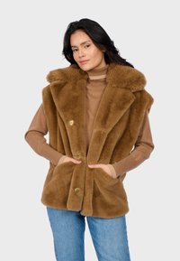 Gilet di pelliccia sintetica marrone con colletto soffice, chiusure con bottoni e tasche laterali. Indossato sopra una maglietta beige, abbinato a jeans blu.