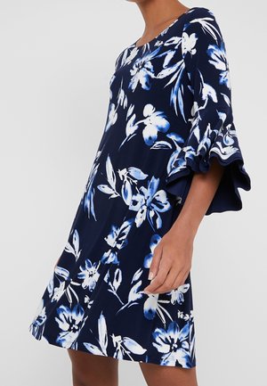 Marineblauwe jurk met witte en lichtblauwe bloemenprint, met brede, gelaagde klokmouwen, gedragen door een persoon die voor een effen achtergrond staat.