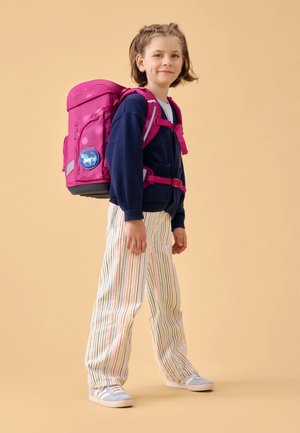 Pink backpack with adjustable straps, two front pockets, and reflective accents. Striped pants and gray sneakers worn by a child.