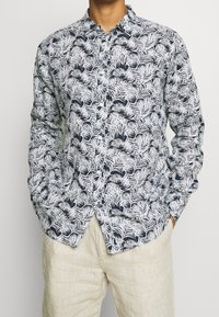 Uomo che indossa una camicia button-up blu navy con motivo a foglie bianche e pantaloni di lino beige, mani nelle tasche, sullo sfondo semplice.