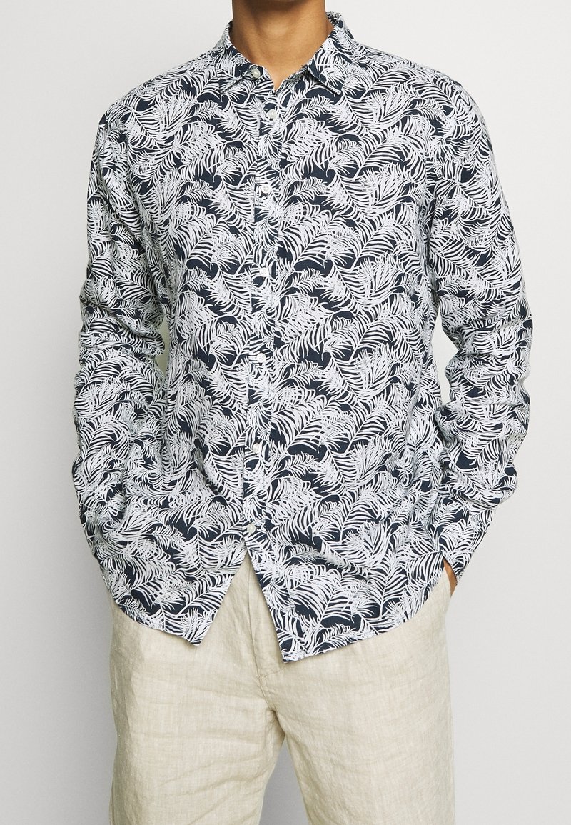 Uomo che indossa una camicia button-up blu navy con motivo a foglie bianche e pantaloni di lino beige, mani nelle tasche, sullo sfondo semplice.
