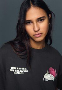 Sötétszürke rövid ujjú póló, bal oldalon fehér szöveggel és rózsaszín virágmintával. Az anyag puha és sima hatású.