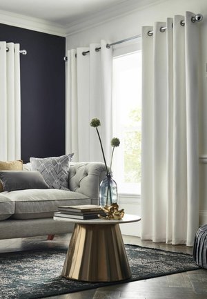 Graues, bezogenes Sofa mit gemusterten Kissen neben weißen Vorhängen und Fenster, runder goldener Couchtisch mit Büchern und Vase mit Blumen auf dunklem Teppich.