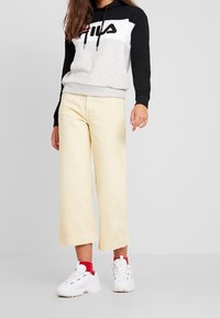 Sudadera con capucha gris y negra con logo rojo y blanco, combinada con pantalones de pierna ancha en amarillo pálido y zapatillas blancas con detalles rojos.