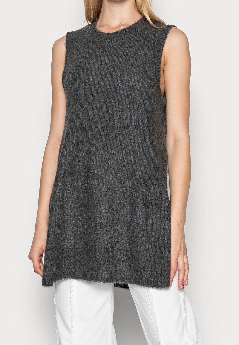 Femme portant un pull-tunique sans manches en maille côtelée gris foncé sur un pantalon blanc, debout devant un fond clair uni.