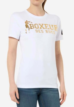 Donna che indossa una maglietta bianca con grafica dorata di un pugile e scritto "BOXEUR DES RUES", abbinata a jeans blu, su sfondo semplice.