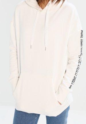 Sweat à capuche blanc avec poche avant et cordons, avec texte noir courant verticalement sur la manche droite, porté avec un jean bleu.