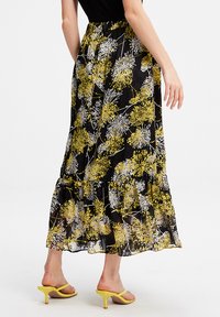 Jupe maxi avec un fond noir ornée de motifs floraux jaunes et blancs, ourlet à volants en plusieurs niveaux et taille élastique, associée à des sandales jaunes.
