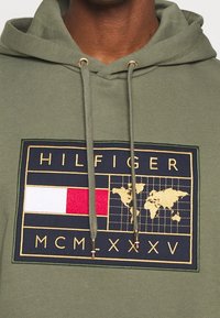 Felpa verde oliva con coulisse, caratterizzata da un grande patch ricamato che mostra un logo, una mappa del mondo e l'anno "MCMXXV" in oro.