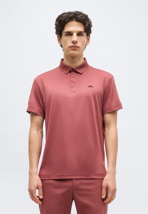 Νεαρός άνδρας με σγουρά μαλλιά, φορώντας ένα ριγωτό πορτοκαλί πόλο με κουμπιά και κοντά μανίκια, καθώς και παντελόνι που ταιριάζει, στέκεται μπροστά σε απλό φόντο.