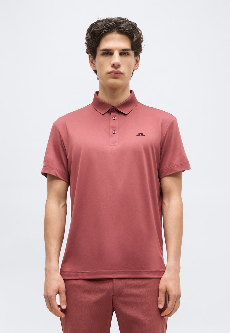 Νεαρός άνδρας με σγουρά μαλλιά, φορώντας ένα ριγωτό πορτοκαλί πόλο με κουμπιά και κοντά μανίκια, καθώς και παντελόνι που ταιριάζει, στέκεται μπροστά σε απλό φόντο.