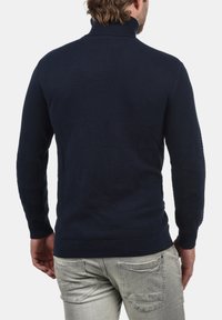Donkerblauwe gebreide trui met een hoge kraag, een gestructureerd patroon en geribbelde boorden, weergegeven van achteren, gedragen met een grijze broek.