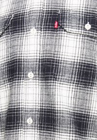 Chemise en flanelle à carreaux noir, blanc et gris. Dispose de deux poches poitrine, de boutons blancs et d'une étiquette logo rouge sur la poche gauche.