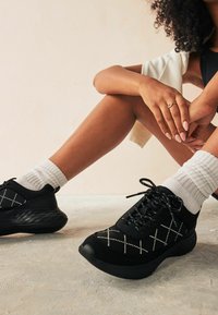 Black athletic sneakers with textured surfaces, featuring white crisscross stitching and laces, set against a soft, neutral background.