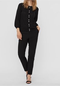 Blusa nera in tessuto strutturato, chiusura a bottone sul davanti e maniche a tre quarti, abbinata a pantaloni neri sartoriali e sandali.