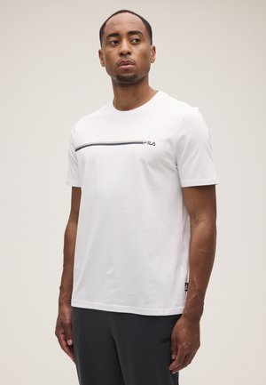 Uomo con una t-shirt bianca a maniche corte FILA con una striscia orizzontale e pantaloni scuri, in piedi e che guarda di lato.