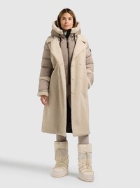 Beige lang frakke med shearling over en tyk polstret jakke, kombineret med cremefarvede sne støvler med snørebånd og en tekstureret overflade.
