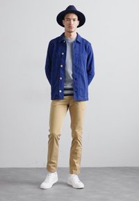 Blue jacket with pockets, gray sweatshirt, beige pants, and white sneakers. Black hat accentuates the outfit. Smooth texture, modern silhouette.