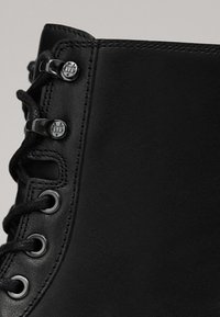 Gros plan sur la tige d'une botte en cuir noir avec des lacets noirs passés à travers des œillets et crochets métalliques, présentant des coutures détaillées.