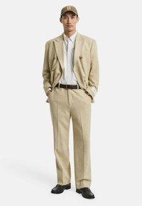 Homme portant un costume beige à double boutonnage avec une chemise blanche, une ceinture marron, des mocassins foncés et une casquette beige avec un logo "G" jaune, les mains dans les poches.