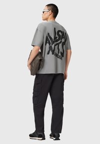 Camiseta gris de gran tamaño con gran texto negro en la parte posterior, combinada con pantalones cargo negros y zapatos deportivos negros. Sosteniendo una chaqueta acolchada.