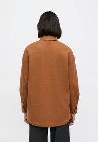 Ruskea ylisuurikokoinen paita, joka on valmistettu pehmeästä kankaasta. Siinä on kaulus, nappikiinnitteiset hihansuut ja kaareva helma. Yksivärinen, ilman kuvioita.