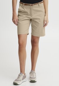 Beige shorts laget av lett materiale, med flat front, sidelommer og glatt finish, sammen med lysegrå sneakers.