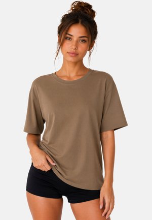 Vrouw met haar in een knot, gekleed in een losse bruine T-shirt en zwarte korte broek, staand tegen een effen lichte achtergrond.