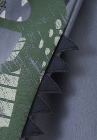 Green dinosaur-themed fabric featuring jagged black accents, on a smooth blue background. Textured print with visible brush strokes.