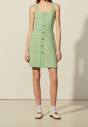 Bezrekawna sukienka w zielono-białą pepitkę z guzikami z przodu, noszona z białymi skarpetkami i kremowymi mokasynami na stojącej modelce.