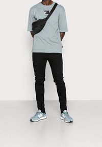 T-shirt bleu clair à manches courtes avec impression graphique, sac bandoulière noir, jeans slim noirs et baskets bleu clair avec des accents gris.