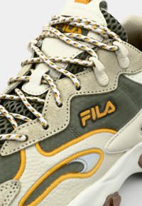 Zapatillas de color gris claro y verde oliva con cuero texturizado, detalles de malla y acentos naranjas. Presentan cordones blancos gruesos y un destacado logo "FILA".