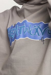Acercamiento de una sudadera de color marrón claro con el texto gráfico azul "HEATWAVE" en el pecho, en una fuente estilizada.