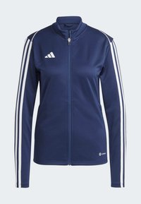 Marineblaue Zip-Jacke mit hohem Kragen, weißen vertikalen Streifen an den Ärmeln und einem kleinen Logo auf der Brust. Weicher Stoff.