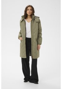 Olīvu zaļas krāsas ilgā volēta jaka ar rāvējslēdzēju priekšpusē, lielu kapuci un sānu kabatām. Valkāta pāri baltai kreklai un melnām biksēm.