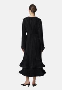 Robe noire plissée avec manches longues, col haut, ourlet à volants et ceinture en tissu. Le matériau texturé crée des lignes verticales et un mouvement fluide.