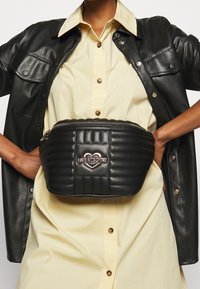 Černá prošívaná ledvinka se stříbrným srdcovým logem, vybavená zipovým uzávěrem a hladkou koženou texturou, nošená přes světle žluté tričko.