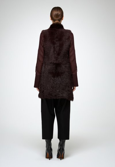 Donna con coda di cavallo castana che indossa un gilet di pelliccia marrone scuro, una maglietta trasparente a maniche lunghe, pantaloni neri cropped e stivali neri con tacco alto, in piedi di spalle.
