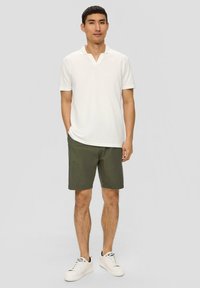 Witte polo met korte mouwen en een V-hals, gecombineerd met olijfgroene shorts. Model draagt witte sneakers en staat tegen een neutrale achtergrond.