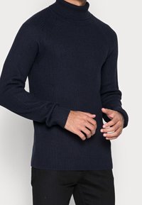 Man som bär en marinblå ribbad turtlenecktröja med långa ärmar och svarta byxor, visad från halsen till höfterna.