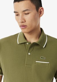 Polo in cotone texturizzato verde oliva, con colletto bianco, due bottoni e logo ricamato sulla tasca del petto.