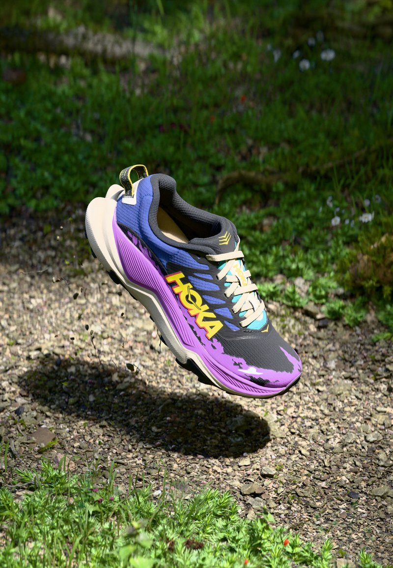 Zapatilla de running Hoka con parte superior en negro y púrpura, superficie texturizada, acentos coloridos y suela acolchada, suspendida sobre grava y hierba.