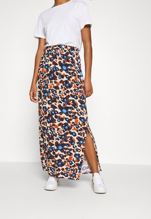 Femme portant un T-shirt blanc, une jupe longue à imprimé floral multicolore avec une fente latérale, et des baskets blanches, debout devant un fond uni.