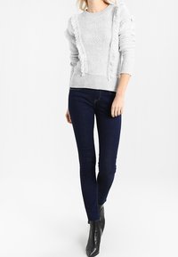 Jasnoszary sweter dzianinowy o teksturowanym wzorze i postrzępionych detalach, w zestawie z ciemnoniebieskimi obcisłymi dżinsami i czarnymi botkami za kostkę.