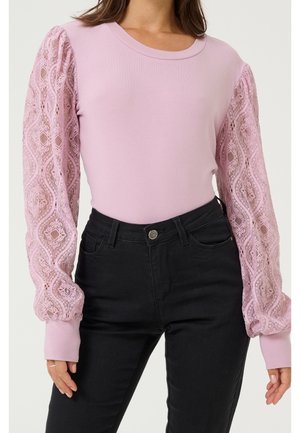 Vrouw draagt een lichtroze top met lange, pofmouwen met kantpatroon en zwarte, hooggetailleerde jeans, staand tegen een effen achtergrond.