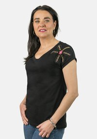 Camiseta de algodón negra con escote en V, mangas cortas y un colorido diseño floral bordado en el hombro izquierdo. Lleva puesta con jeans azules.