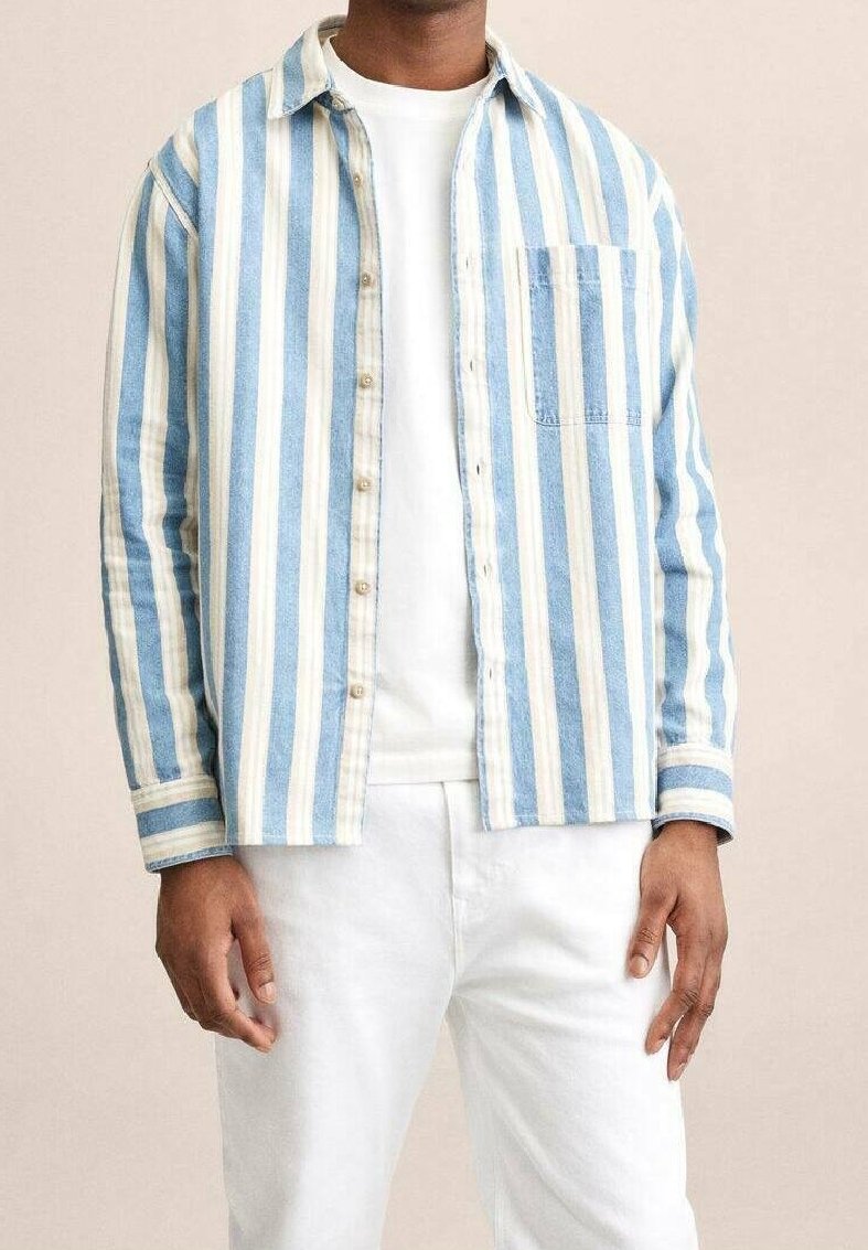 Homme portant une chemise à boutons bleu clair et blanche à rayures verticales sur un T-shirt blanc uni et un pantalon blanc, debout devant un fond beige.