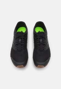 Een paar zwarte mesh hardloopschoenen met zwarte veters, fluorescentgroene binnenzolen en bruine rubberen teenvakken, gezien van bovenaf op een witte achtergrond.