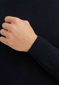 Pull en marine foncé avec des poignets côtelés, montrant la zone des manches et une main partiellement serrée. Le matériau semble doux et tricoté.