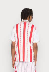 Rot-weißer gestreifter Sporttrikot, kurze Ärmel, mit vertikalen Linien und Pfeilmuster. Mit rundem Kragen und kontrastierenden Akzenten.